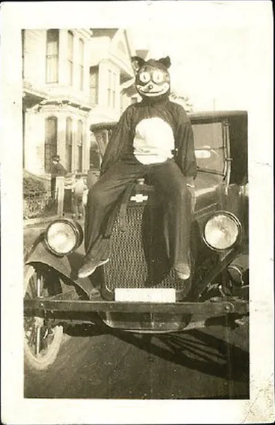 A Halloween reveler in a cat mask sitting on the hood of an automobile, 1920s snapshot