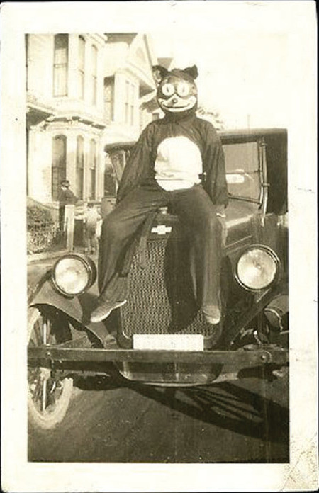 A Halloween reveler in a cat mask sitting on the hood of an automobile, 1920s snapshot, c. 1917 — historic photograph