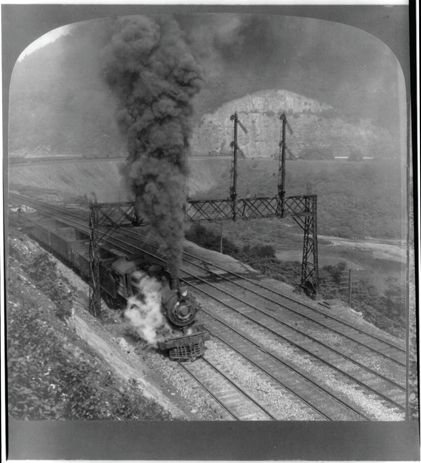 Steam locomotive with smoke passing signal gantries beside a rocky cliff — historic image