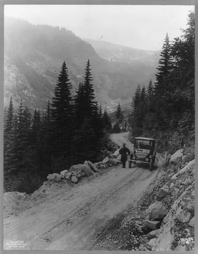 [Detroit Electric" auto on promotional tour through mountains from "Seattle to Mt. Rainier": Man standing in front of auto which is parked on mountain road]"