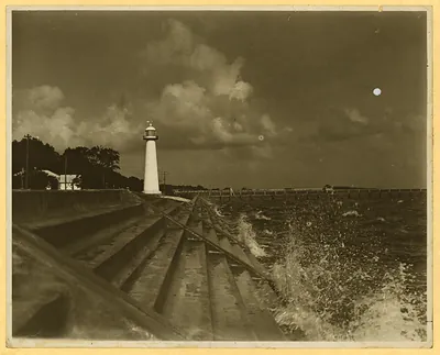 White lighthouse above stepped seawall, waves breaking, sepia photograph