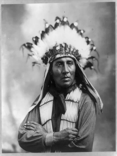 [Running Horse, half-length portrait, facing slightly right with arms folded; wearing feather headdress and bone breast plate]