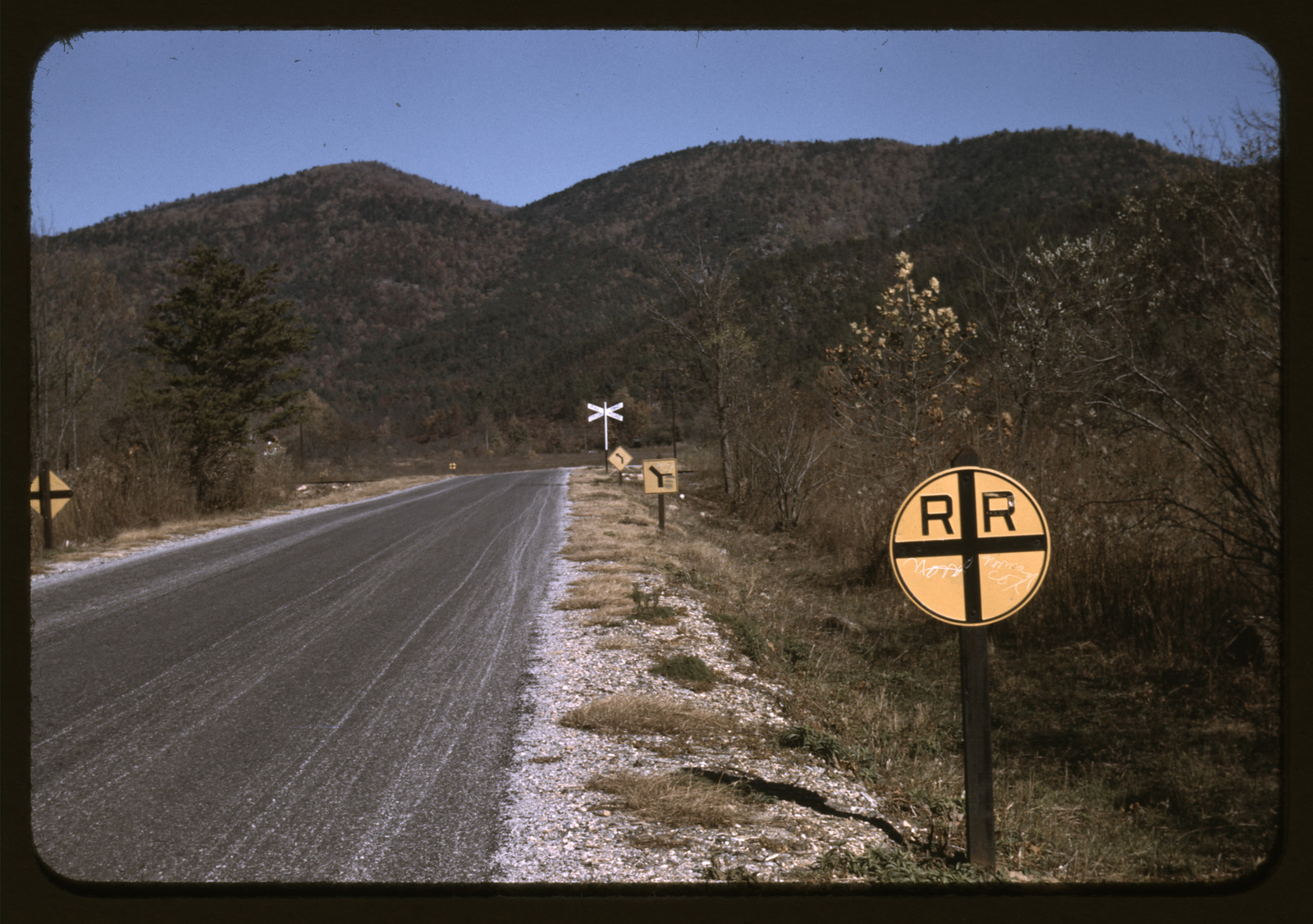 Paved mountain road with railroad crossing signs and wooded hills — historic image