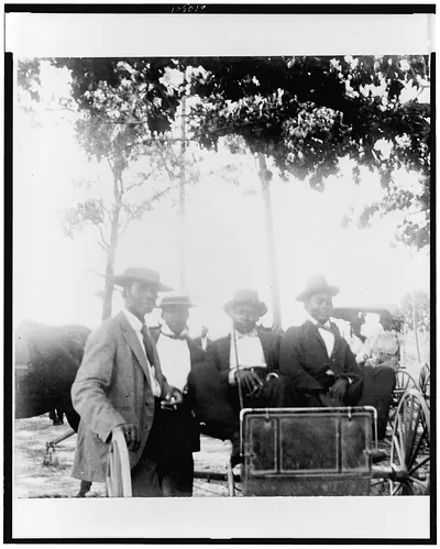 [Two African American men in horse-drawn carriage and two standing alongside]