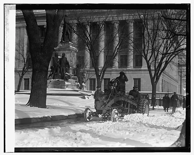 Men clearing deep snow with a machine beside a monument