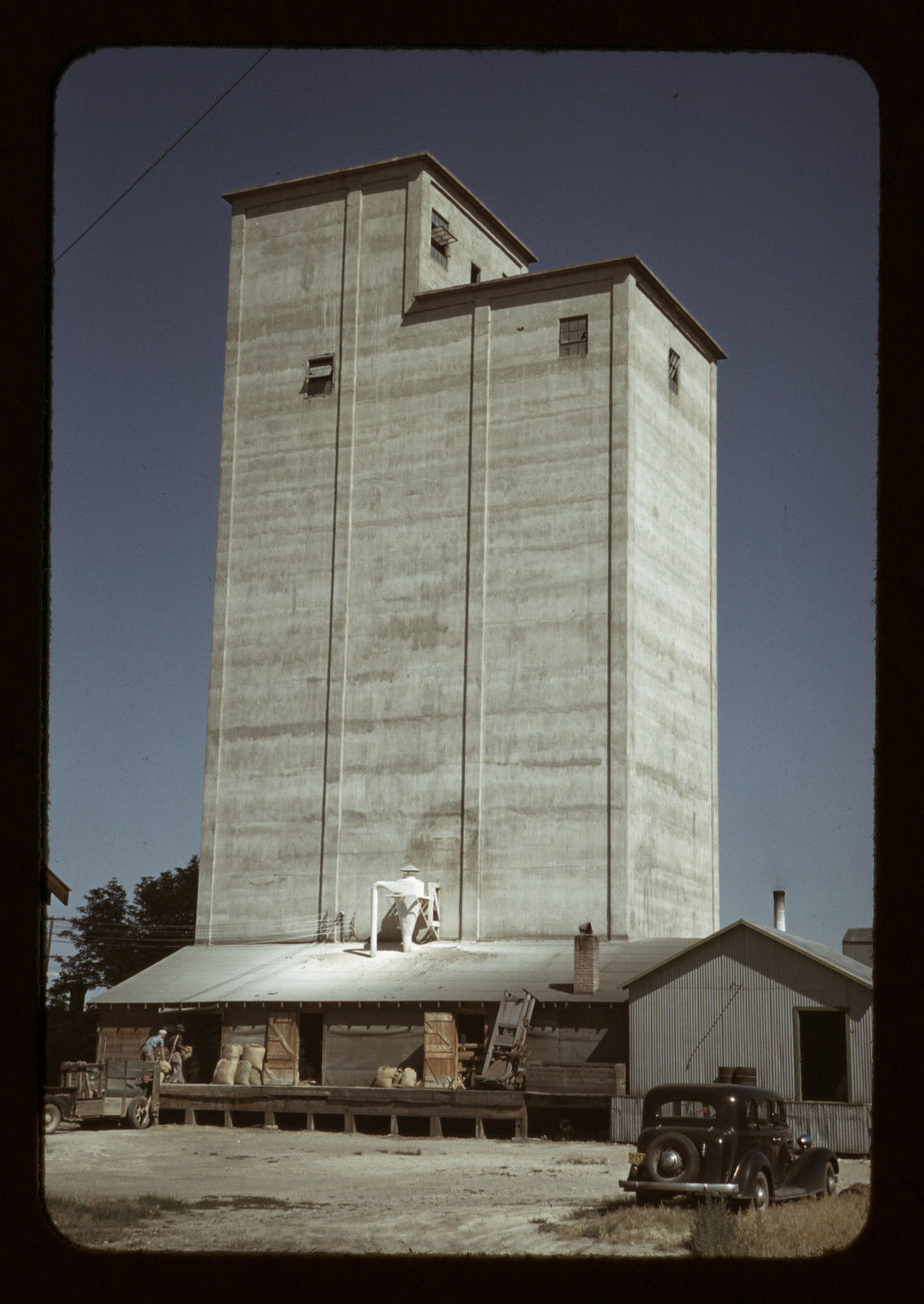 Concrete grain elevator with warehouse and parked vintage car — historic image