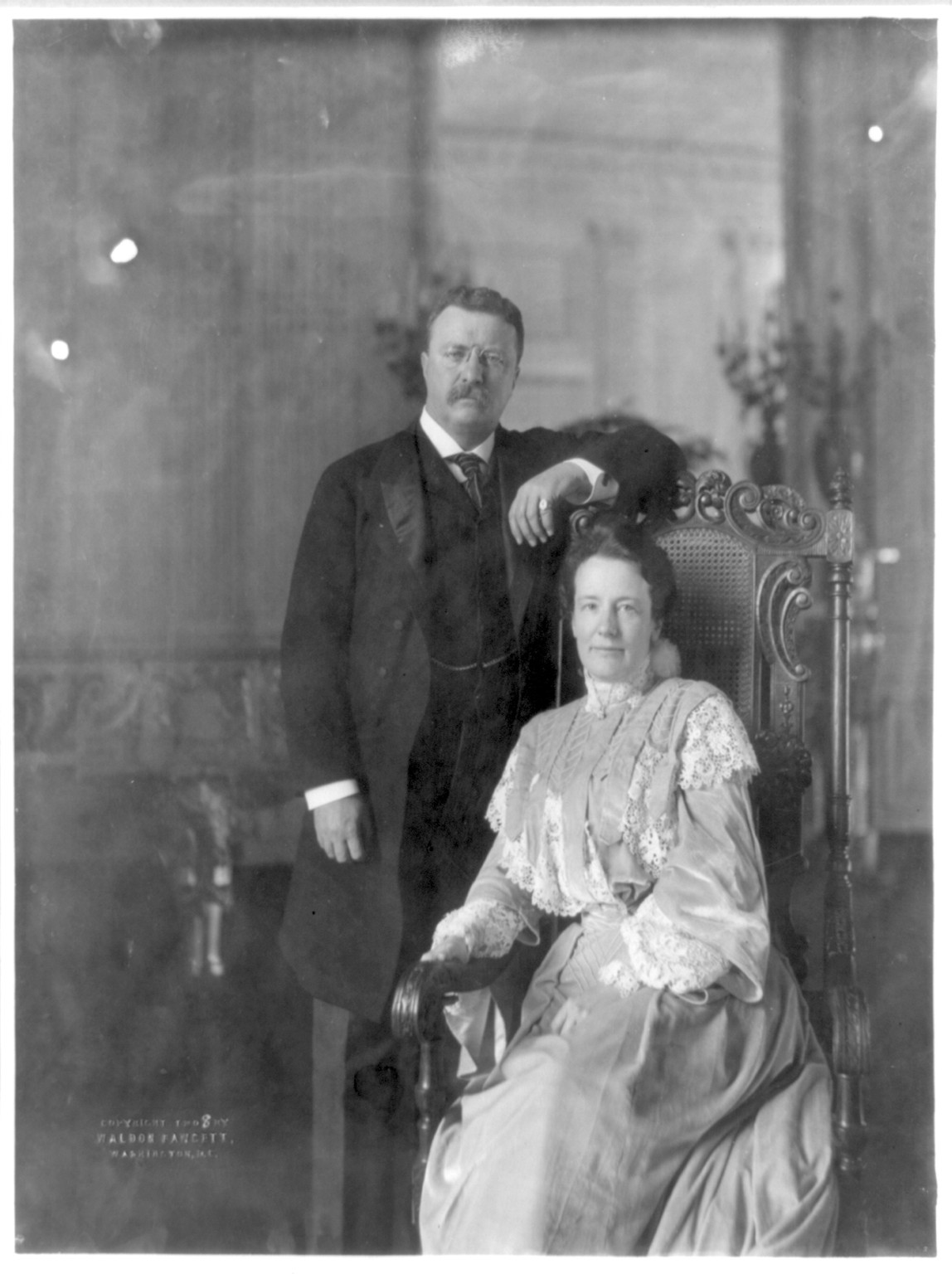 [Theodore Roosevelt, three-quarter length portrait, standing, facing right; alongside his wife, seated], c1908. — historic photograph