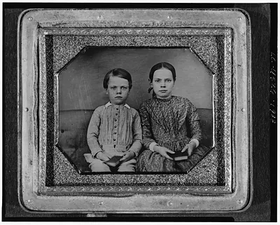 [Group portrait of an unidentified boy and girl, three-quarter length, seated on upholstered bench, facing front, holding books]