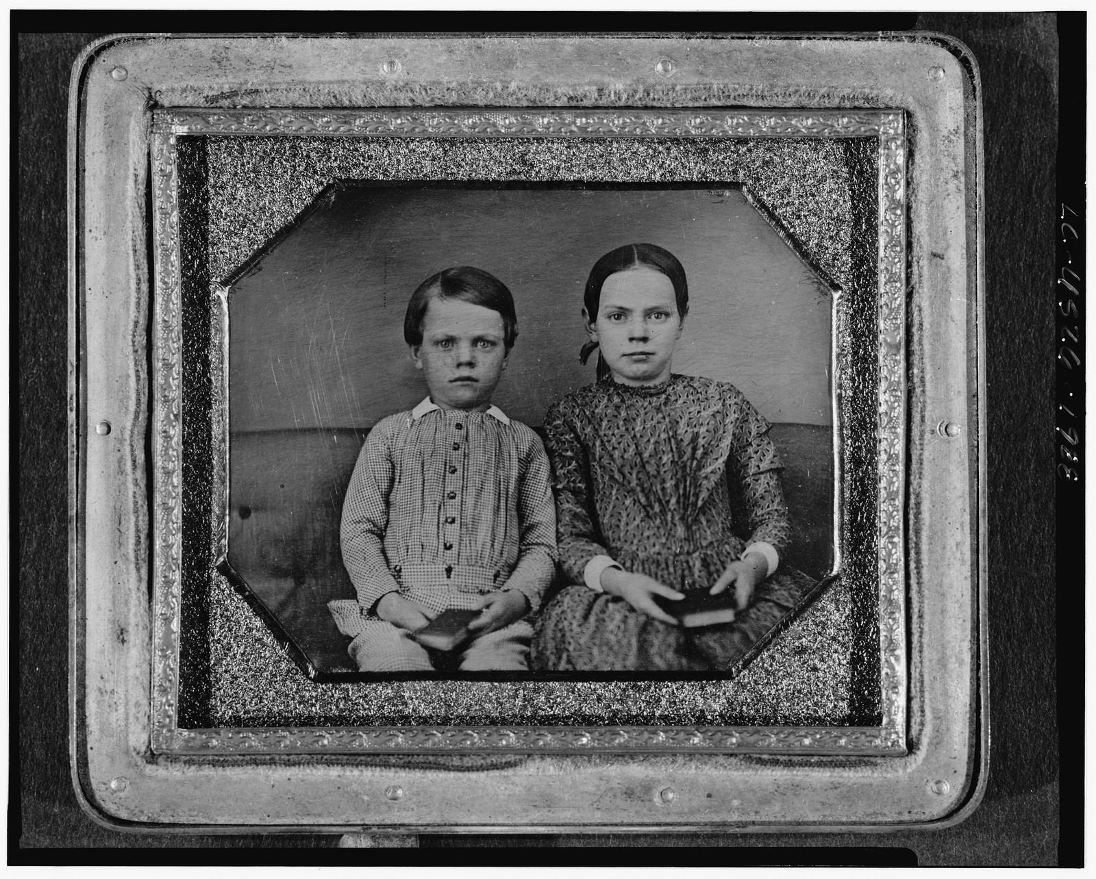 [Group portrait of an unidentified boy and girl, three-quarter length, seated on upholstered bench, facing front, holding books], [between 1840 and 1860] — historic photograph