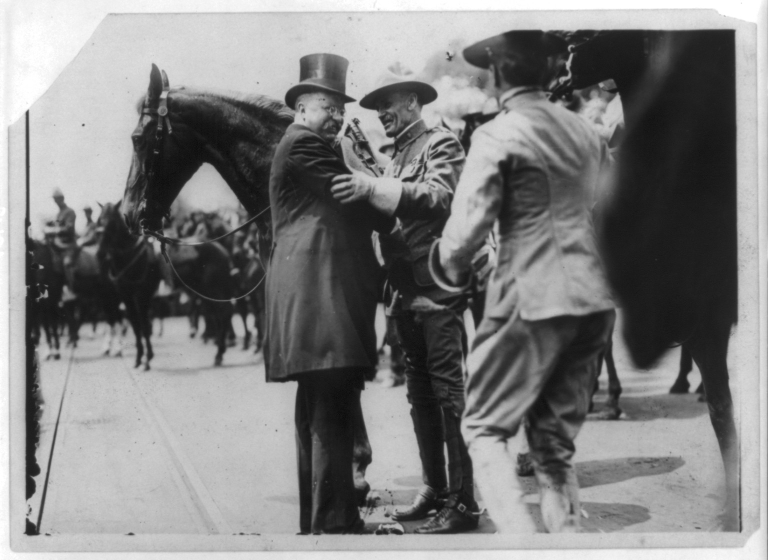 [Theodore Roosevelt, full, standing, facing right, greeting Col. B-, of Rough Riders, in front of horse, with other men on horseback in background], c1910. — historic photograph
