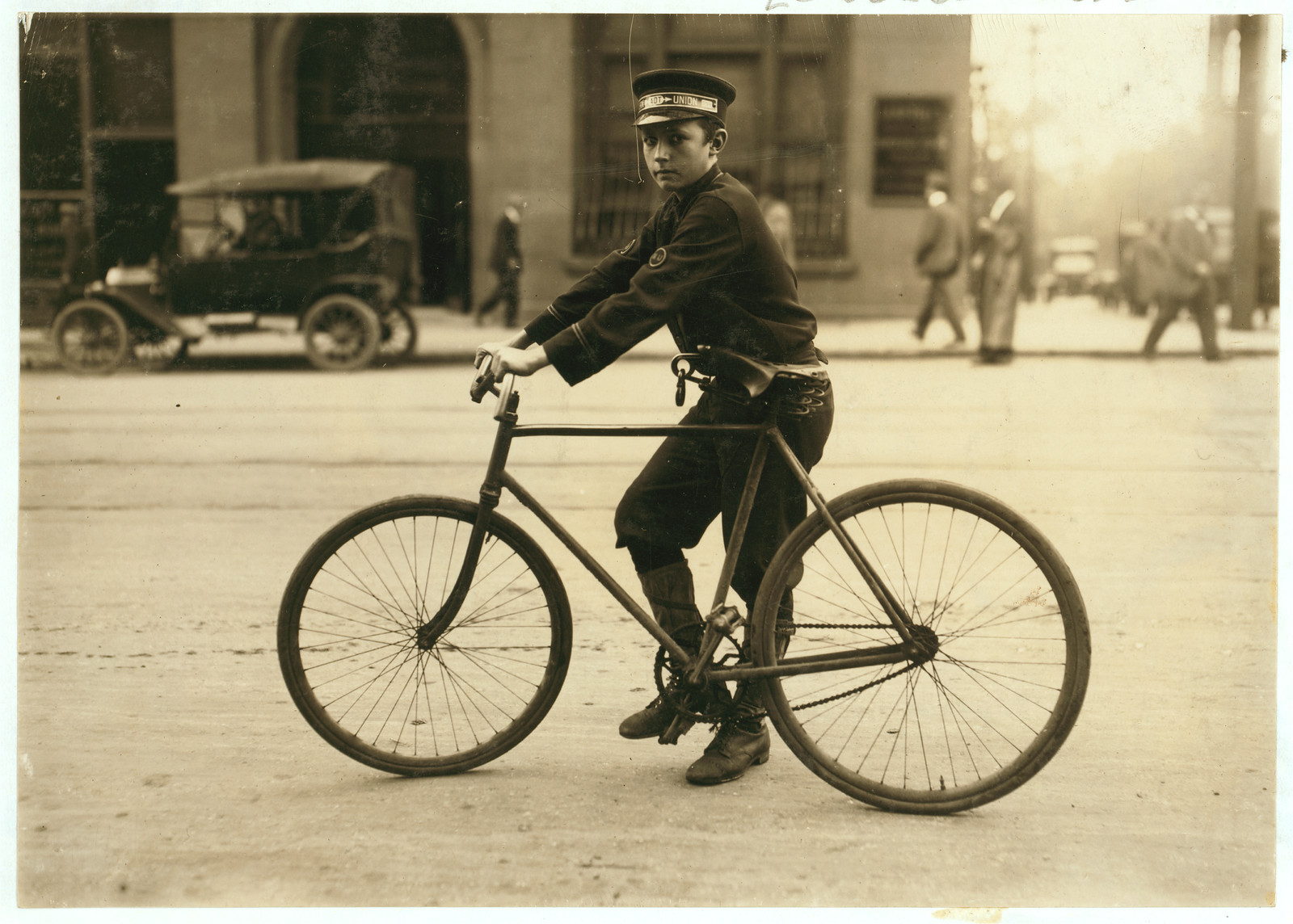 Boy messenger in cap riding a bicycle on city street — historic image