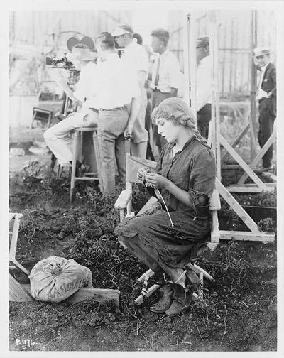 [Mary Pickford knitting a sweater for the American Red Cross, with movie film crew behind her]