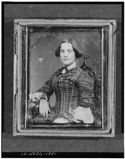 [Unidentified woman, half-length portrait, facing front, seated in chair with arm resting on table with tablecloth]