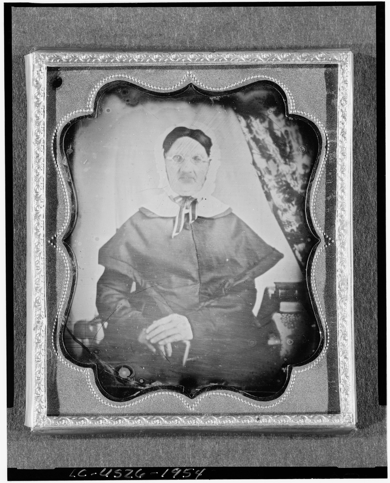 [Unidentified woman with spectacles, seated beside table with tablecloth, with books and curtain to right], [between 1850 and 1860] — historic photograph