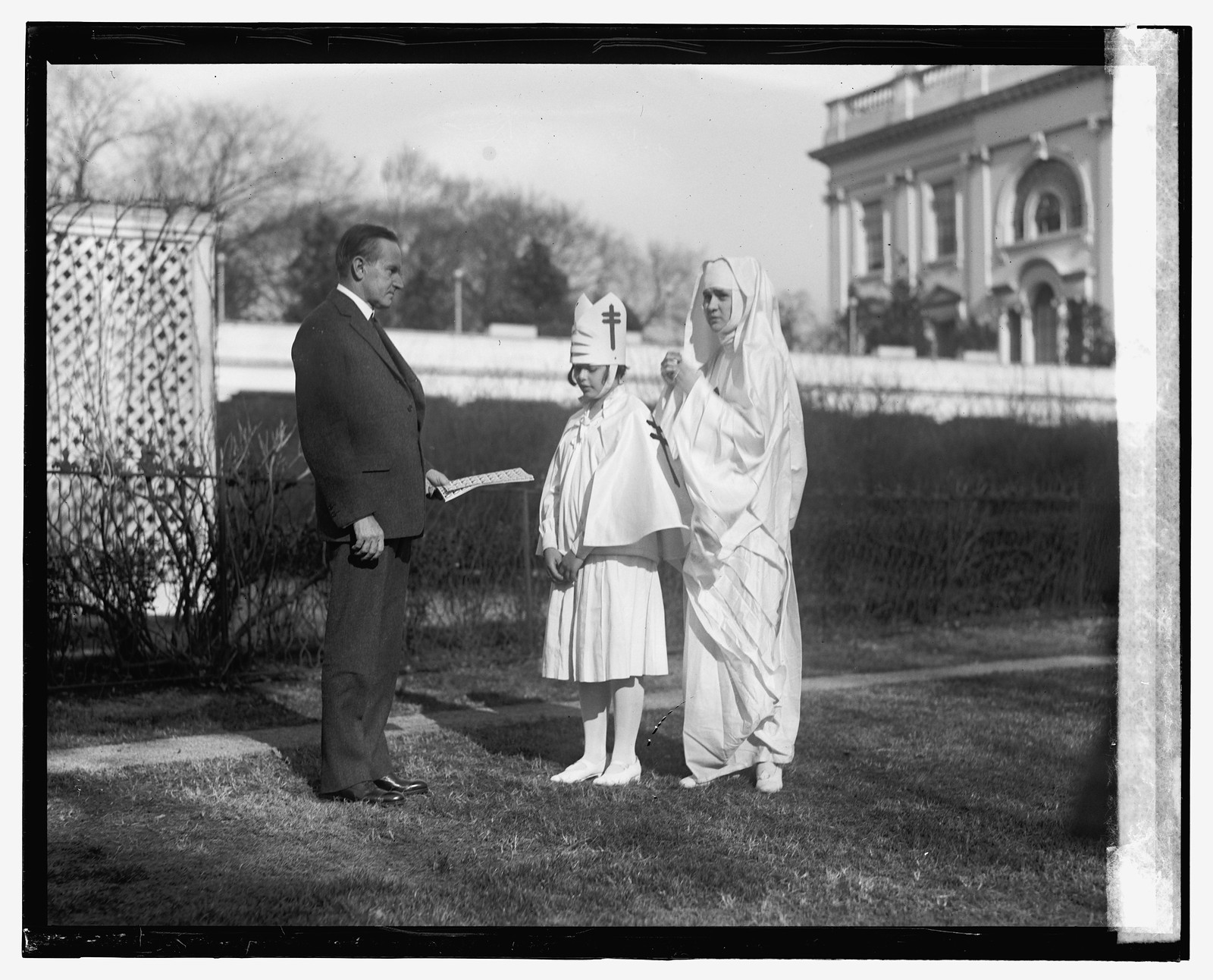 Man in suit with a child in a white robe and hat, and a robed figure, outdoors — historic image