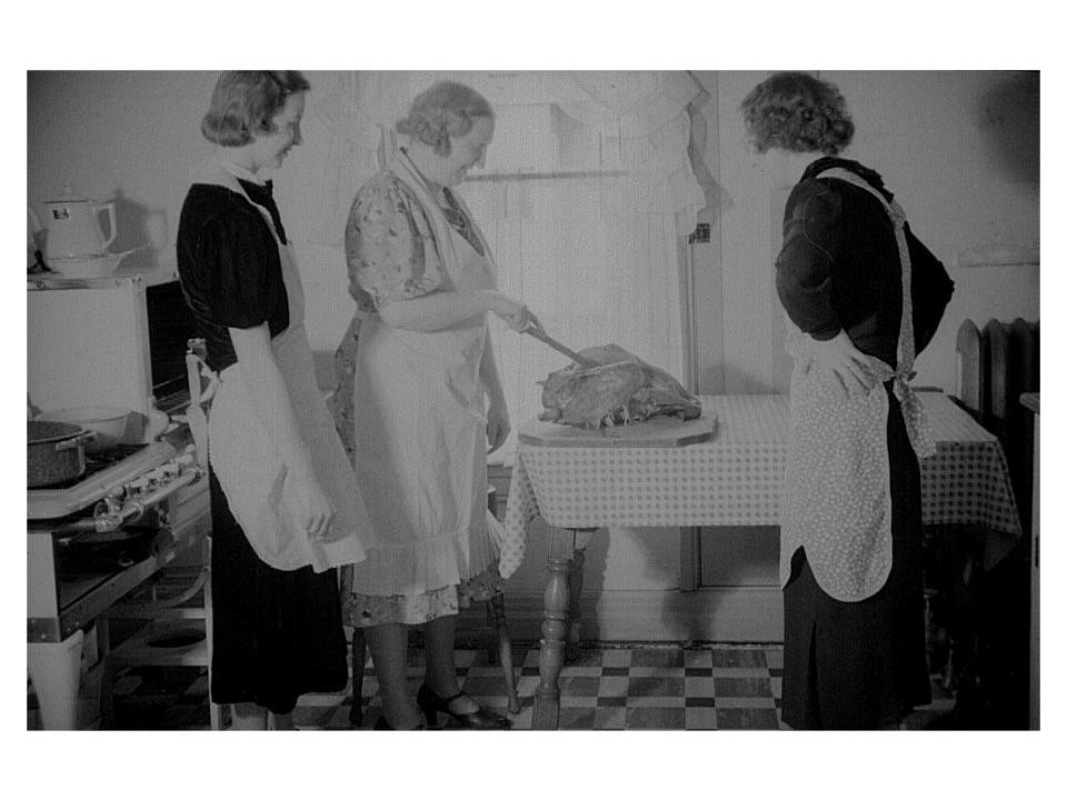Three women in a kitchen, one carving a roast bird — historic image