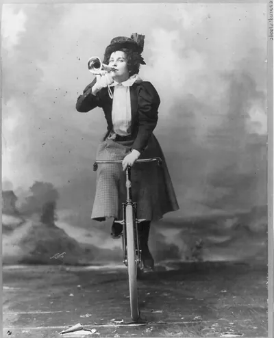 [Madge Lessing, full length, on bicycle, facing left; holding musical horn to lips]