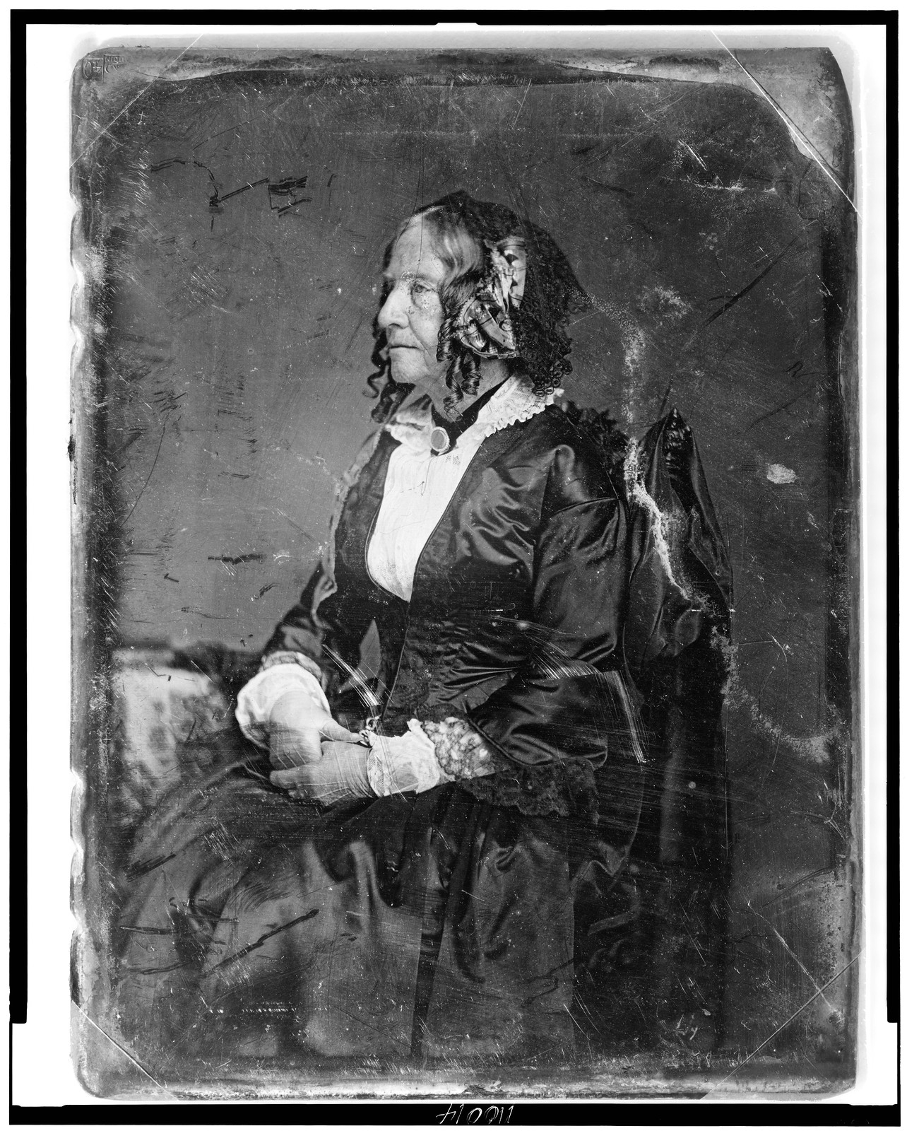 [Unidentified woman, about 65 years of age, three-quarter length portrait, nearly profile to the left, seated in chair by table, hands in lap, wearing cap and light gloves], [between 1850 and 1860] — historic photograph