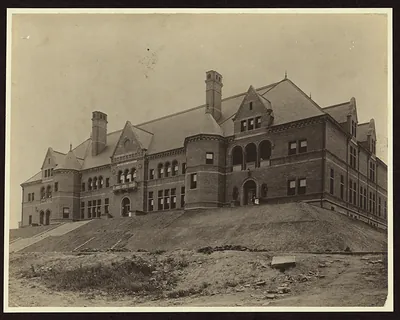 Large brick public building on a grassy hill, sepia photograph
