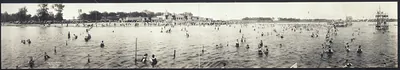 Crowded lakeside beach with swimmers and pavilion, panoramic photograph