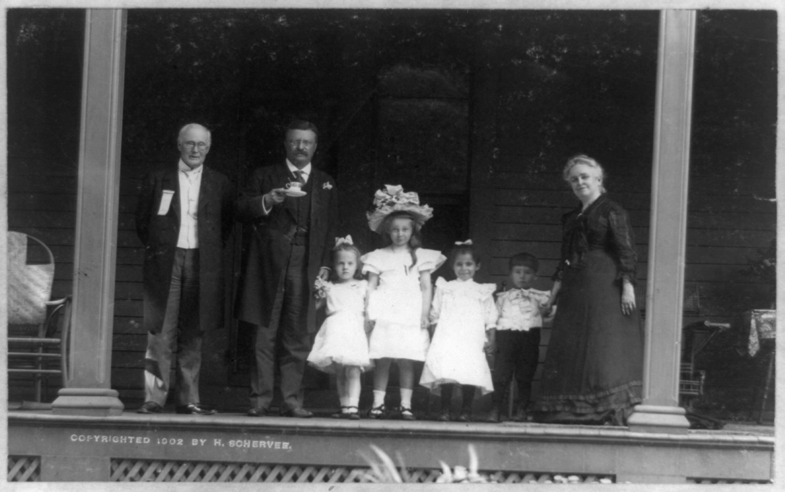 [Theodore Roosevelt, full-length portrait, standing on porch, facing front, holding cup and saucer, with older man, older woman, and four children], c1902. — historic photograph