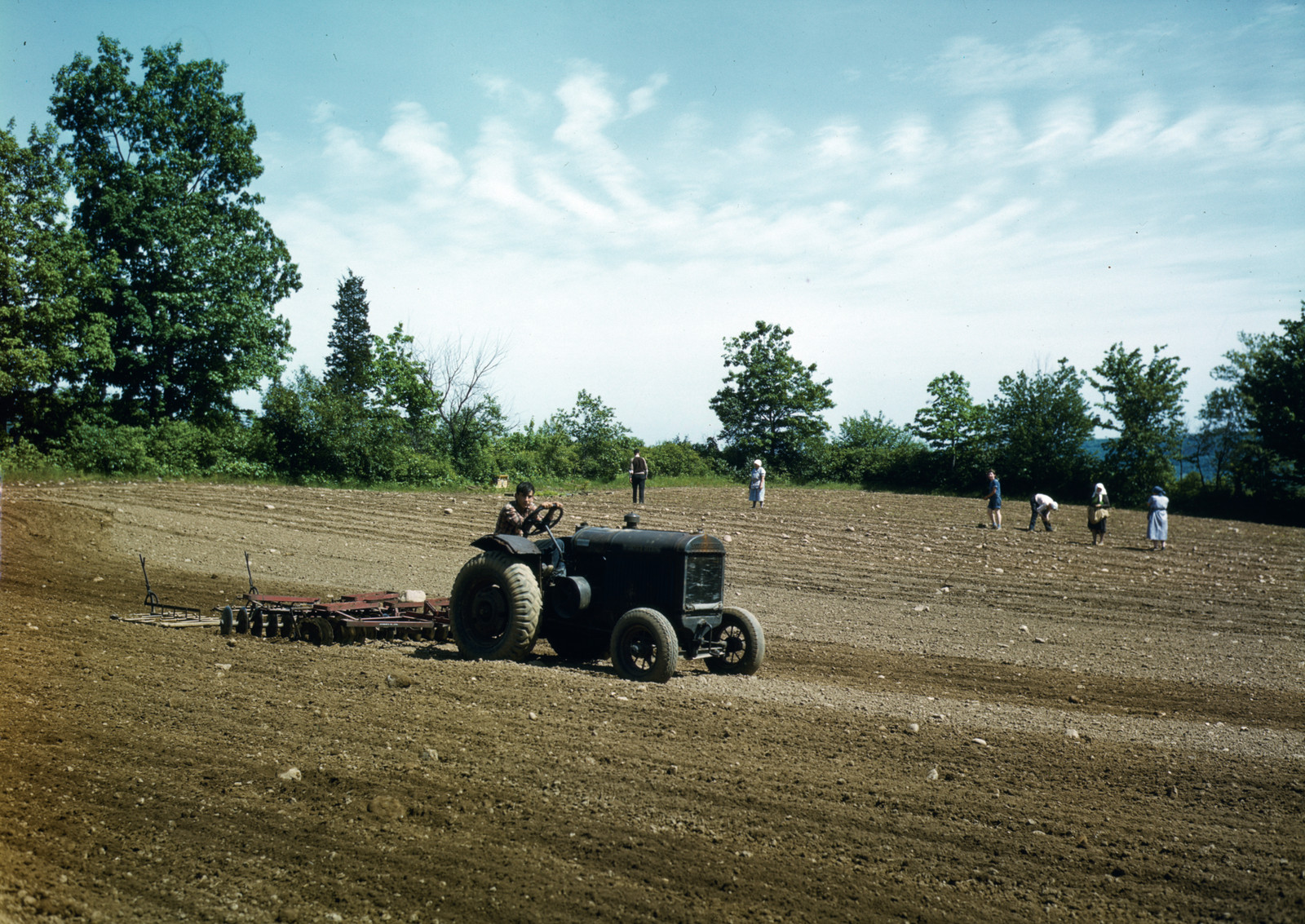 Tractor pulling a disc harrow across a field with workers — historic image