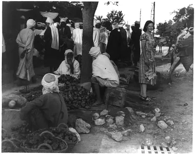 [Woman, wearing western dress, standing among tomato vendors at outdoor market in North Africa]
