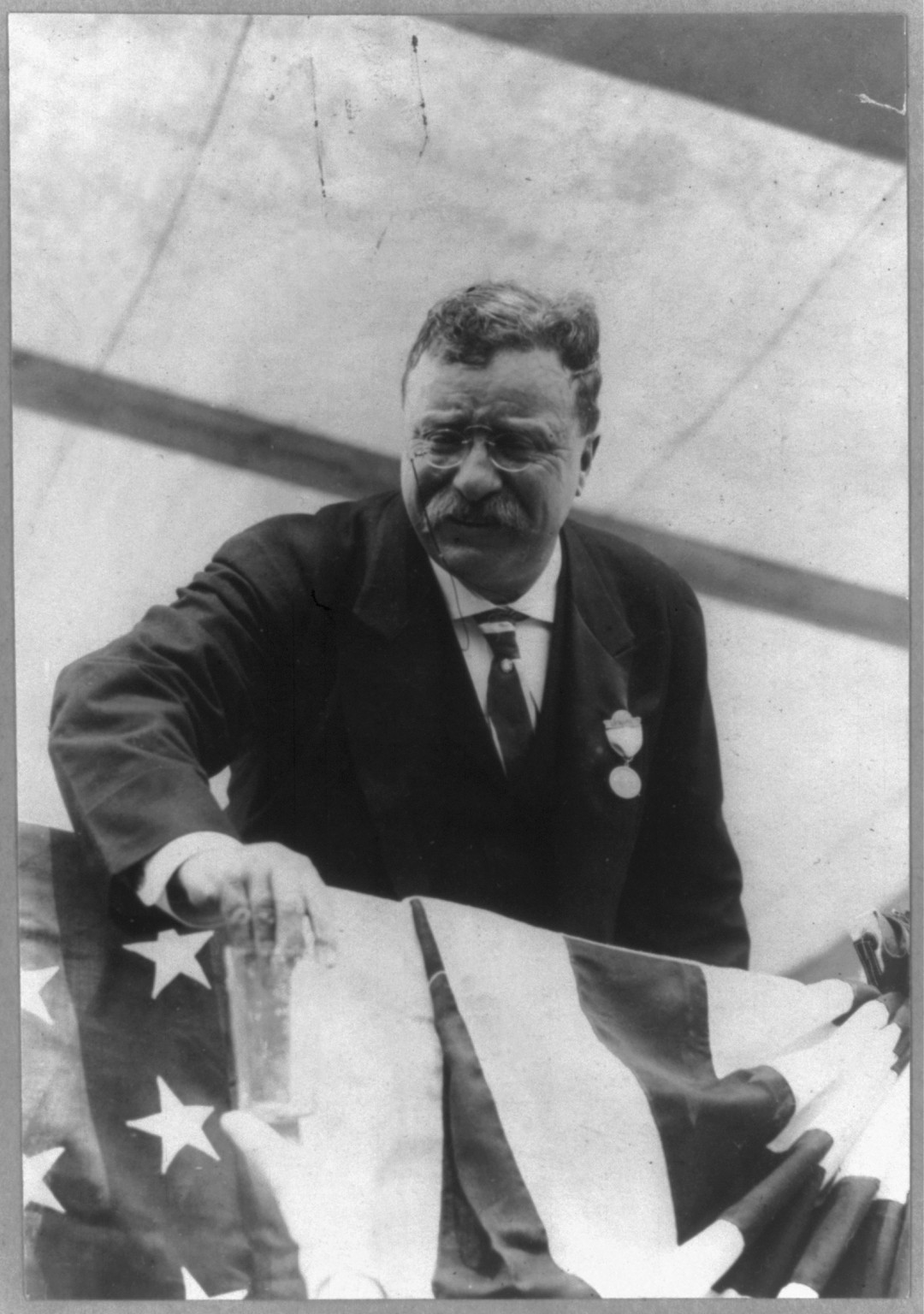 [Theodore Roosevelt, half-length, standing, U.S. Flag draped in front, facing left], c1910 June 27. — historic photograph