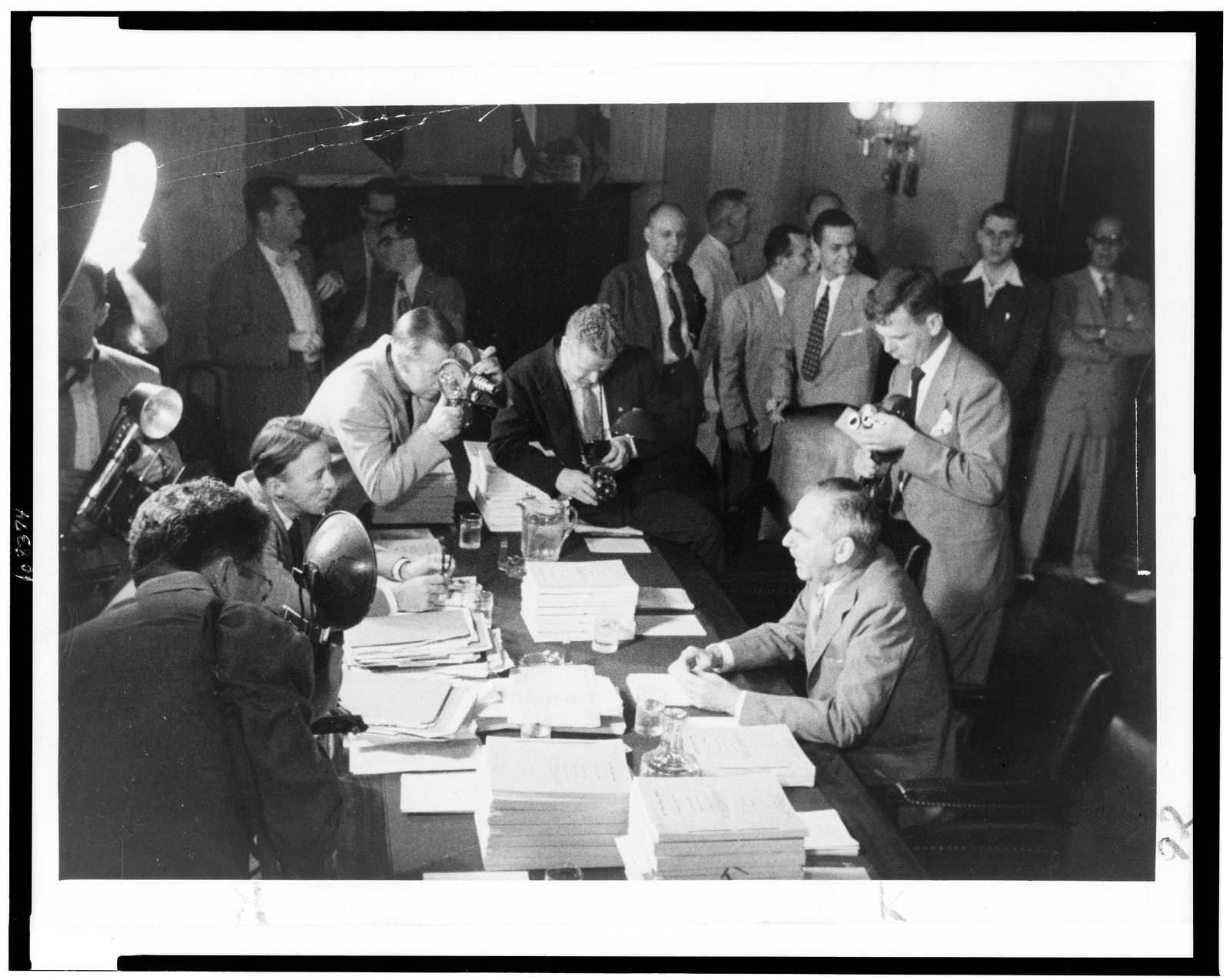 [Dean Acheson, half-length portrait, seated at table, facing left, surrounded by news reporters] — historic photograph