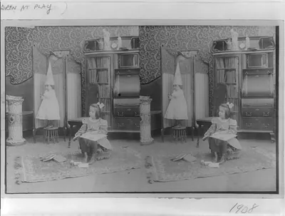[Girl seated in middle of room with books; smaller child standing on stool and wearing dunce cap]