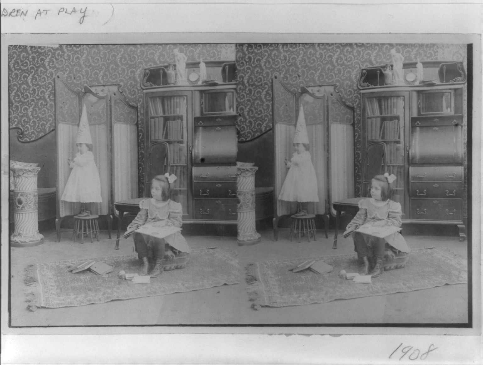 [Girl seated in middle of room with books; smaller child standing on stool and wearing dunce cap], c1908. — historic photograph