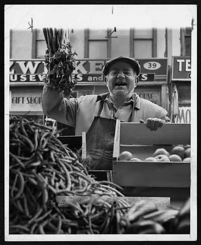 Smiling vegetable vendor holding celery beside crates of apples