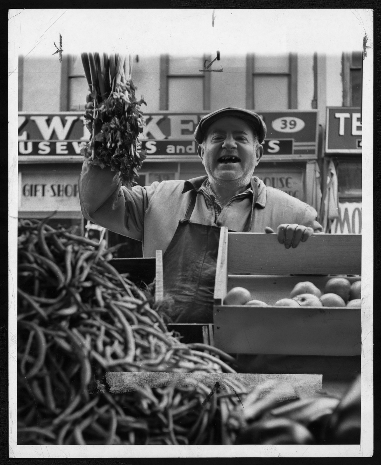 Smiling vegetable vendor holding celery beside crates of apples — historic image