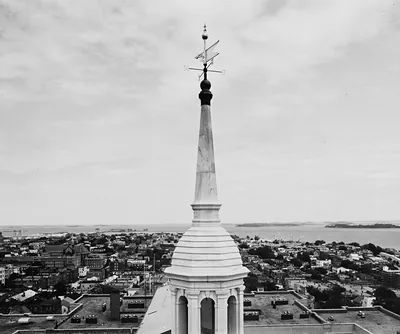Black and white photograph of a steeple with weathervane over a harbor city