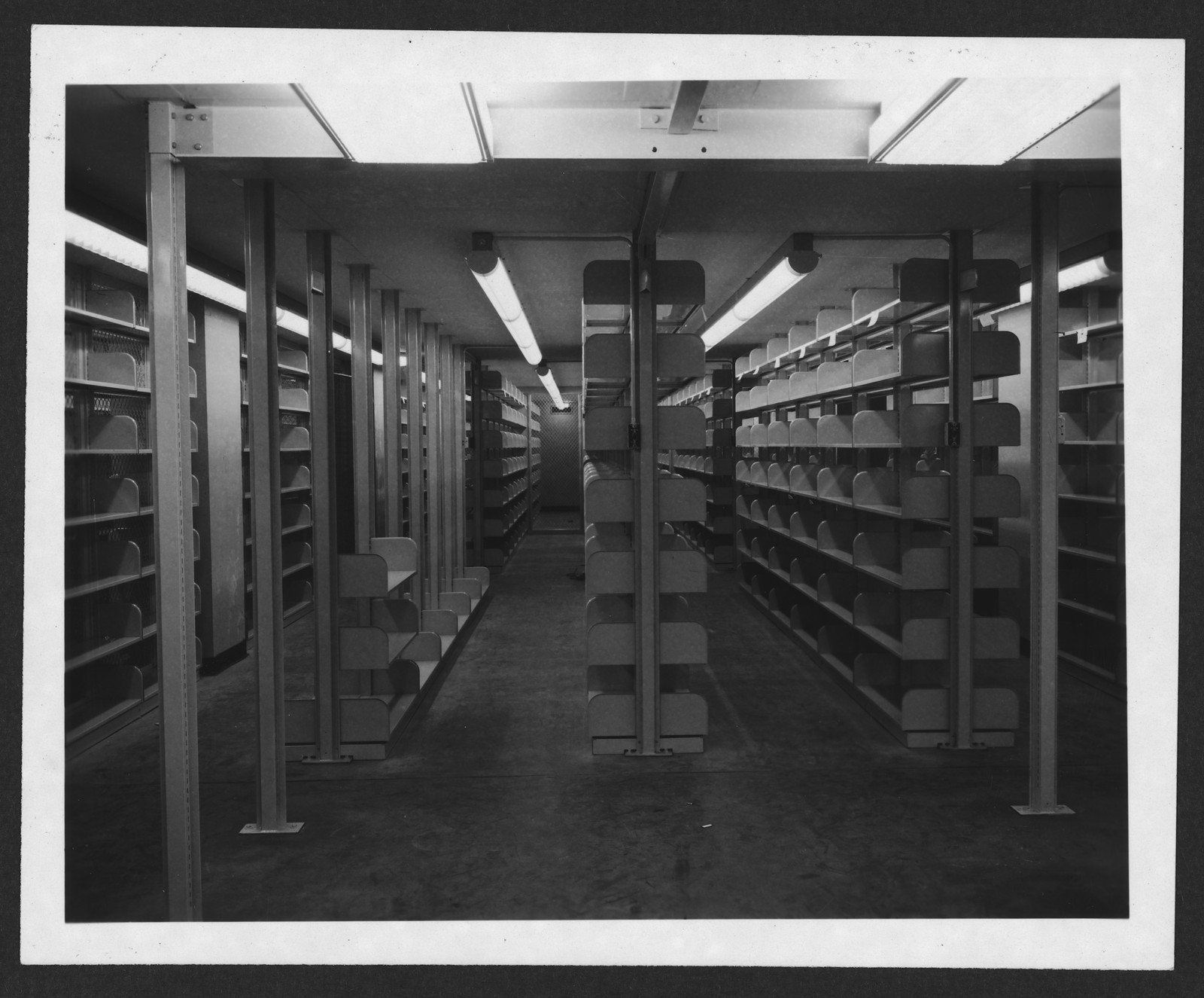 Library book stacks with metal shelving and fluorescent lights — historic image