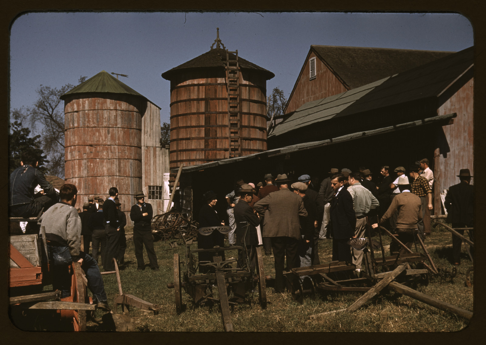 Men gathered at a farm auction beside silos and barn — historic image