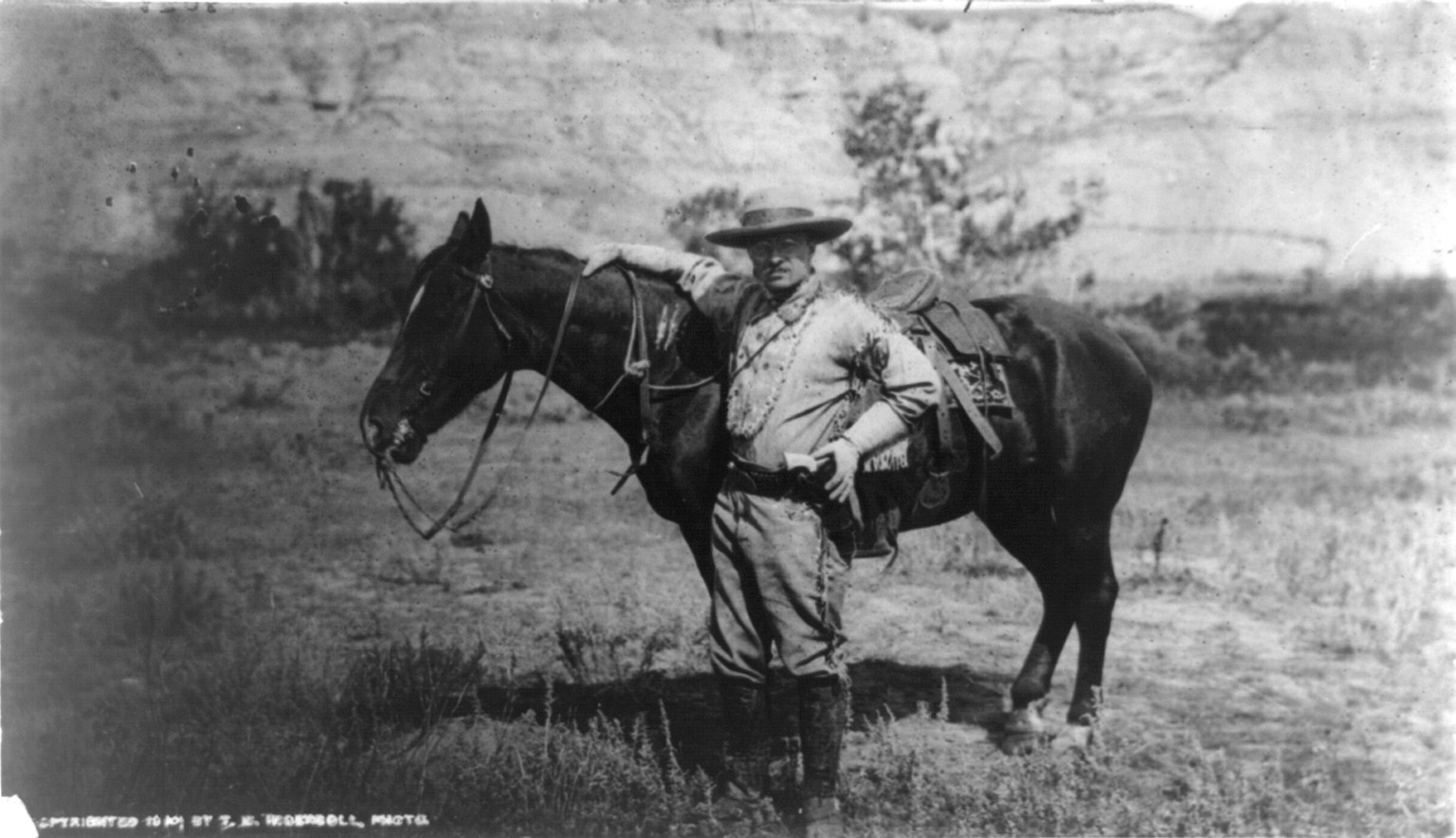 [Theodore Roosevelt, full-length portrait, standing alongside horse, facing left; wearing cowboy outfit], c1910. — historic photograph