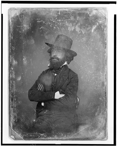 [Unidentified man, three-quarter length portrait, facing left, sitting in chair, wearing hat and a dark glove on one hand]