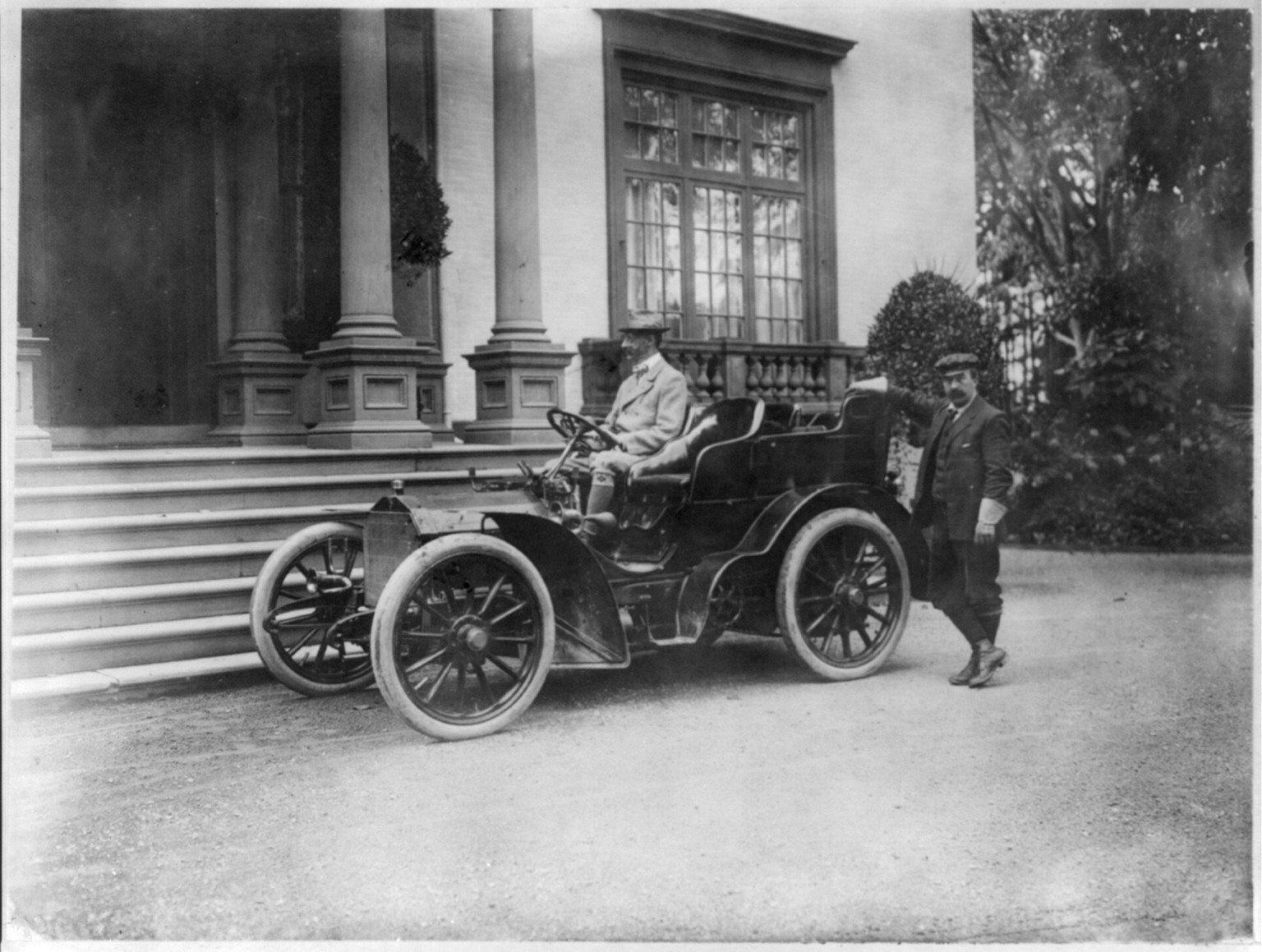 John Jacob Astor in automobile [with chauffeur standing at rear of the automobile], c1903. — historic photograph