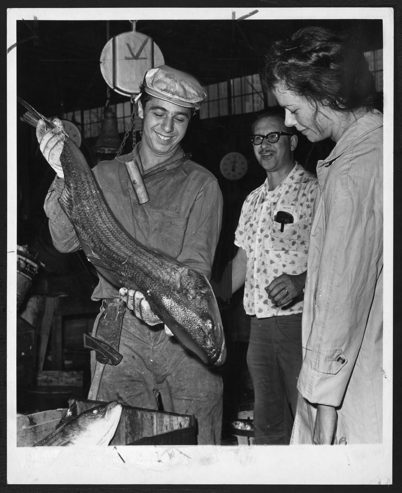 Man in cap showing a large striped bass at market — historic image