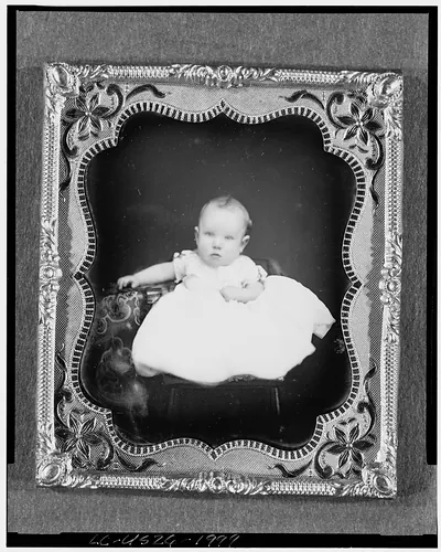 [Harrison Grey Fiske, full-length portrait of six month old baby boy, seated on chair, propped next to table with tablecloth]