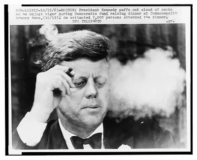 [President John F. Kennedy, head-and-shoulders portrait, smoking a small cigar at a Democratic fundraiser in Boston's Commonwealth Armory]