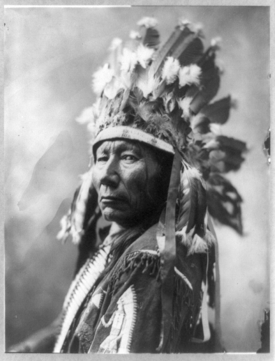[Stands Hard, head-and-shoulders portrait, facing left, in feather headress], c1900. — historic photograph