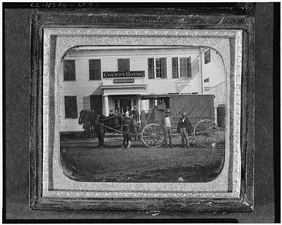 [View of a horse and covered cart in front of white frame building with signs for County House, J. Griggs and L.J. Phillips, Daguerrian Rooms, in Thompson, Connecticut]