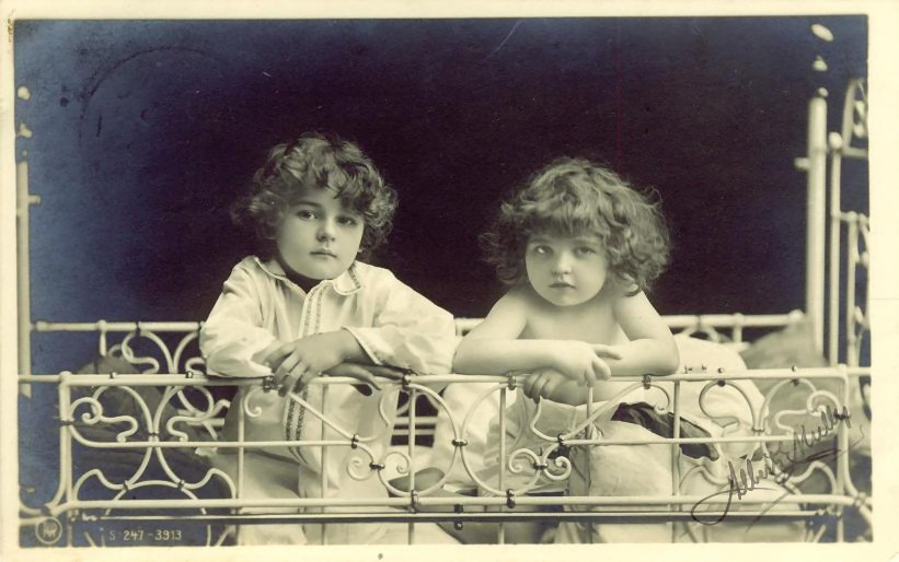 Two curly-haired children behind an ornate white iron bed rail — historic image