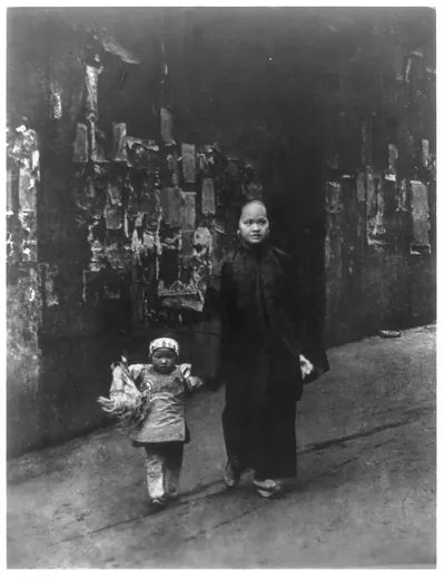 [Mother and small daughter walking on sidewalk in Chinatown. San Francisco. Old ragged and faded handbills on wall in background]