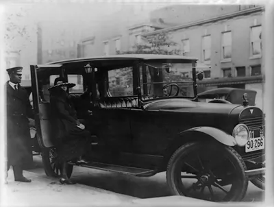 [Woman getting into back seat of car as chauffeur holds door, Washington, D.C., area]