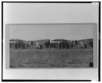 [Group of Native Americans (possibly Winnebago) wearing blankets some standing before log fence others sitting canopied shelter in background]