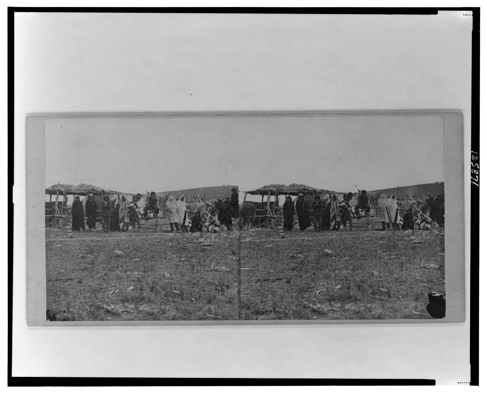 [Group of Native Americans (possibly Winnebago) wearing blankets some standing before log fence others sitting canopied shelter in background], 1862 — historic photograph