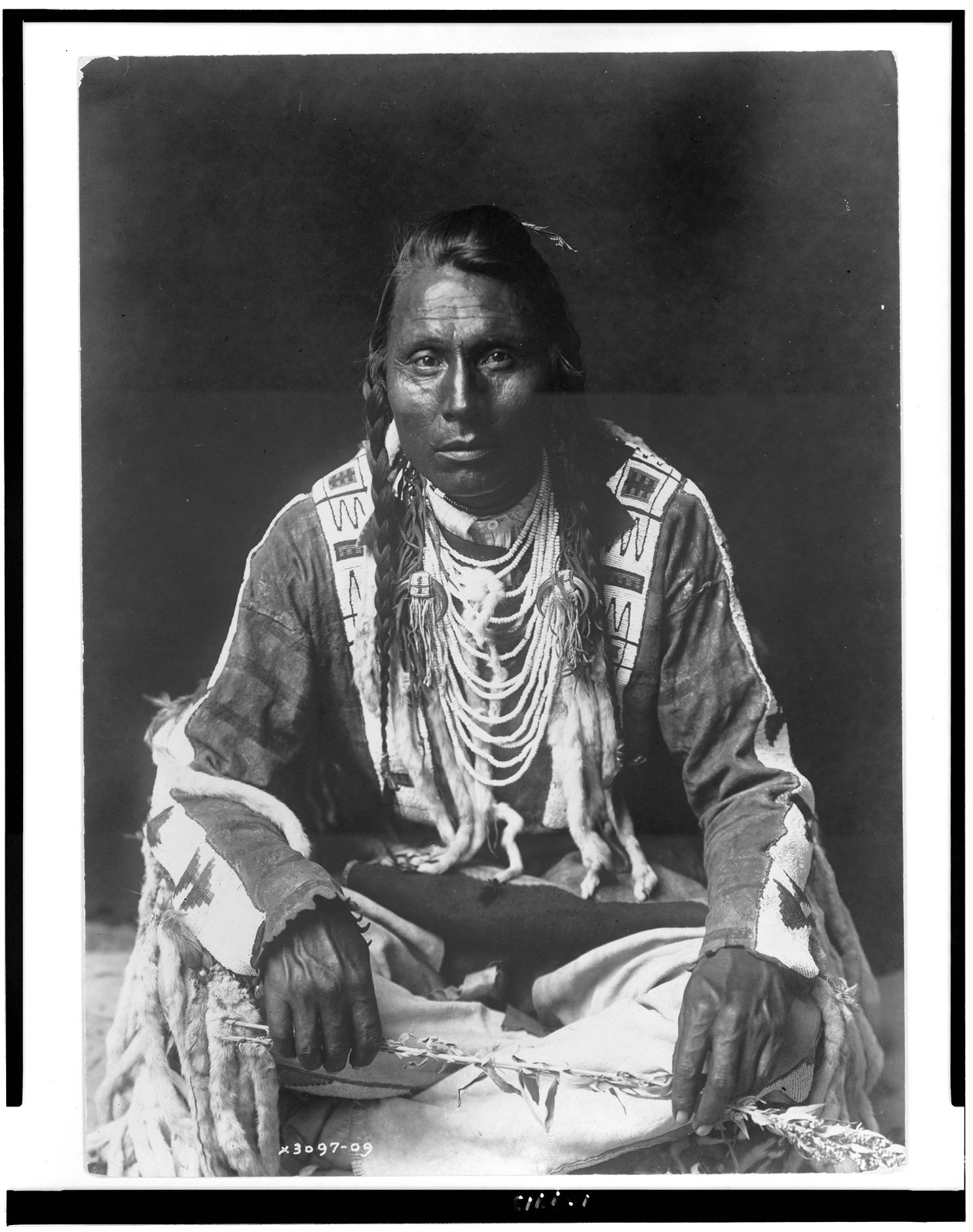 [Wades in Water, Piegan Indian, full-length portrait, seated on floor, facing front, with braids, beaded buckskin shirt and leggings, beads with ermine tail trim], c1910. — historic photograph